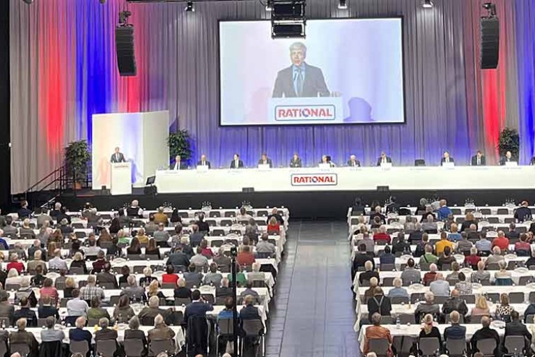A large crowd at a general meeting in a conference hall, with a speaker on stage.