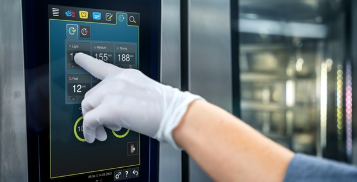 Mano con guante operando la pantalla táctil de un RATIONAL iCombi Pro en una cocina profesional.