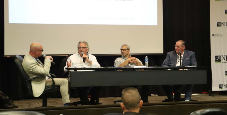 Four men sit on a hospitality panel as one speaks into a microphone beneath a projected presentation.
