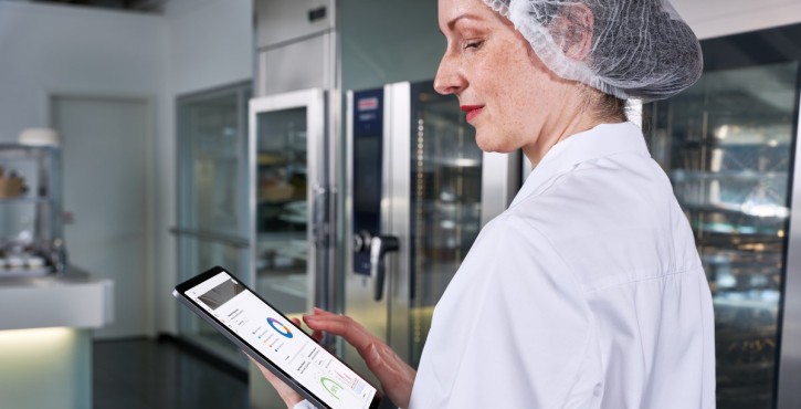 A female chef with a hairnet on walks around the kitchen with HACCP information on her ipad.