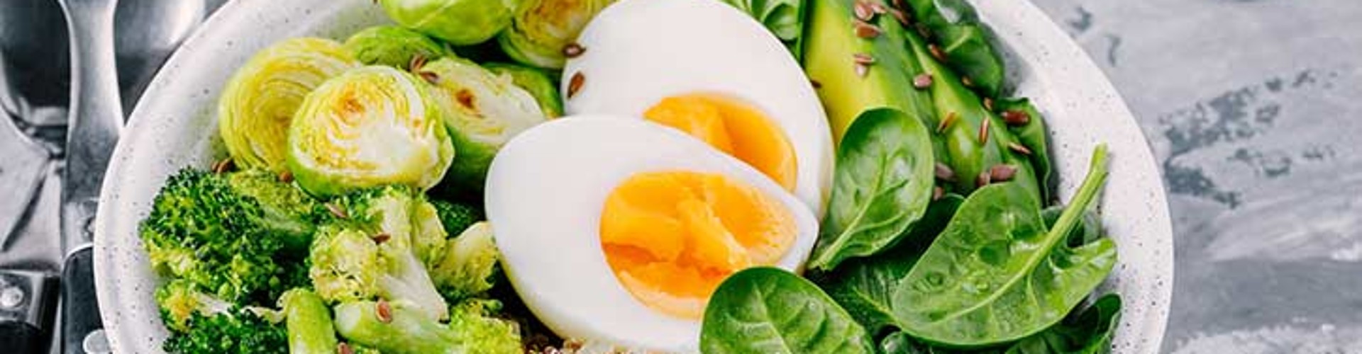 A colorful vegetarian Buddha bowl featuring quinoa, boiled eggs, broccoli, Brussels sprouts, and fresh spinach on a textured surface.