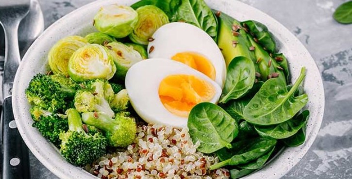 A vegetarian buddha bowl full of spinach, avocado, sprouts, broccoli, hard boiled eggs and quinoa is served in a white bowl.