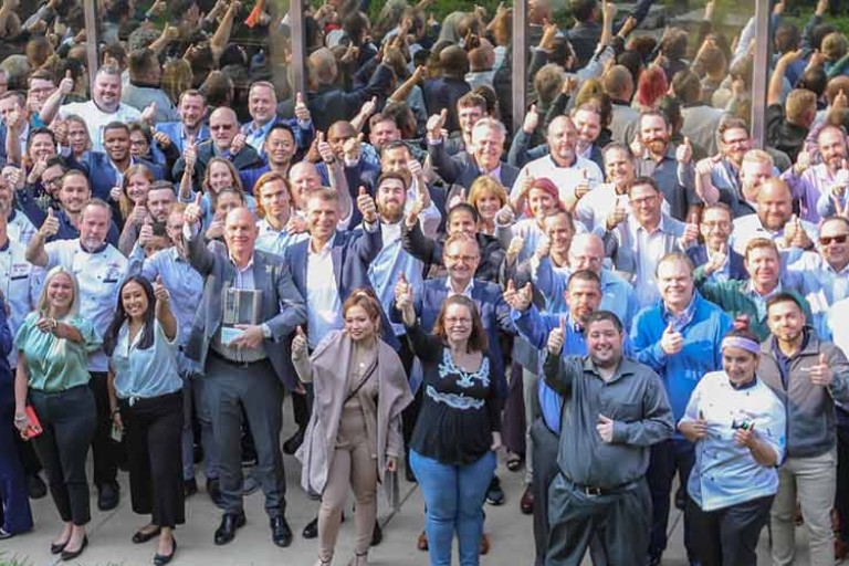 Large group photo of Rational USA employees in business attire and chef uniforms, outdoors, giving thumbs up in front of a glass building.